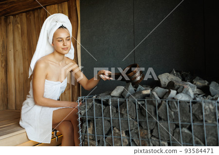 Young woman wrapped in a towel with a wooden ladle pours water on hot stones in the sauna 93584471