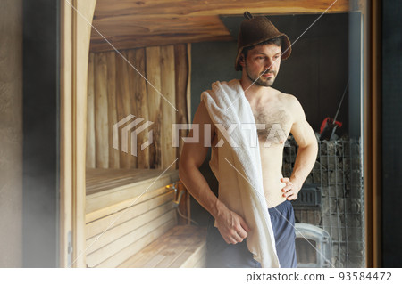 Strong man in a bath hat and a towel on his shoulder stands in the sauna Strong man in a bath hat and a towel on his shoulder stands in the sauna 93584472
