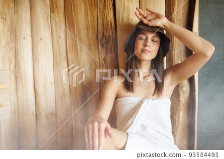 Woman in a bath hat wrapped in a towel relaxes in a hot sauna 93584493