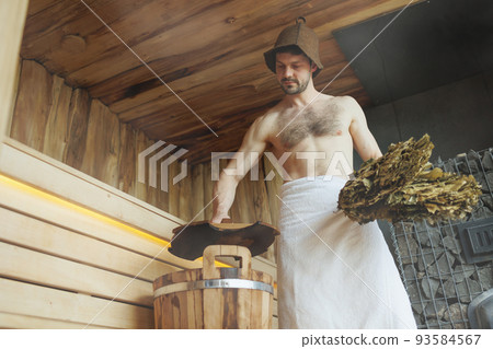 Handsome man with a muscular body in a towel with a bath broom in his hand opens the wooden lid of the bucket in the sauna 93584567