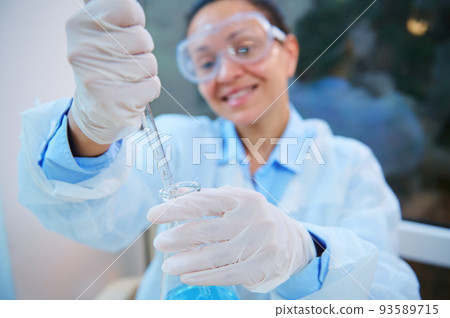 Selective focus on glass dropper in gloved hands of blurred woman scientist chemist wearing safety goggles and lab coat, doing science experiments, pipetting chemical solution in biological laboratory 93589715