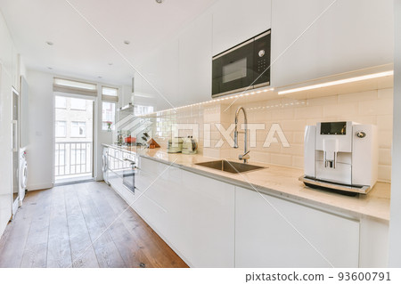 Fragment of interior of light narrow home kitchen with minimalist style white furniture with sink and stove Fragment of interior of light narrow home kitchen with minimalist style white furniture with sink and stove 93600791