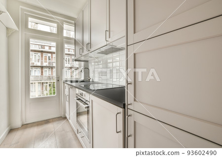 Fragment of interior of light narrow home kitchen with minimalist style white furniture with sink and stove Fragment of interior of light narrow home kitchen with minimalist style white furniture with sink and stove 93602490