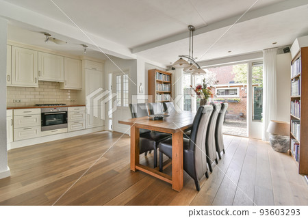 Modern minimalist style interior design of studio apartment with open white kitchen and dining zone with table and chairs illuminated by loft style lamp Modern minimalist style interior design of studio apartment with open white kitchen and dining zone with table and chairs illuminated by loft style lamp 93603293
