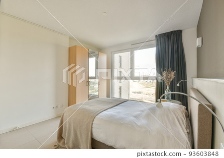 Retro bedside table with books and lamp located near comfortable bed and shelf with decorations against window in modern bedroom 93603848