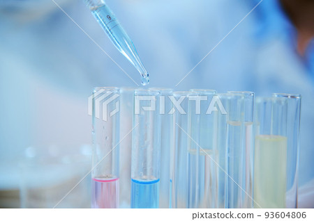Selective focus on a drop of a blue liquid substance dripping from a glass laboratory pipette into a test tube on a tripod. Chemical science experiment or analysis in a clinical research laboratory 93604806