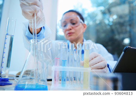 Focus on test tubes with going on chemical reaction on blurred background of a female scientist, lab assistant pipetting liquid substance into flat bottomed flask in scientific research laboratory Focus on test tubes with going on chemical reaction on blurred background of a female scientist, lab assistant pipetting liquid substance into flat bottomed flask in scientific research laboratory 93604813