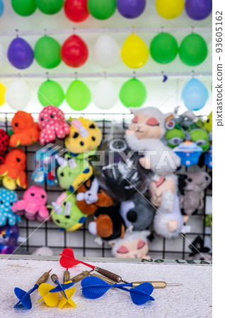 selective focus on darts sitting on a ledge with a wall of balloon targets and prizes that a player can win at a fair. selective focus on darts sitting on a ledge with a wall of balloon targets and prizes that a player can win at a fair. 93605162