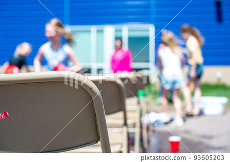 Selective focus on rows of metal folding chairs at an outdoor school function with blurred children in the background. Selective focus on rows of metal folding chairs at an outdoor school function with blurred children in the background. 93605203