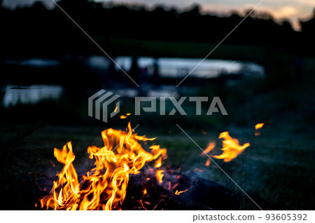Leaping flames of a camp fire with a darkening view of a pond in the background. 93605392