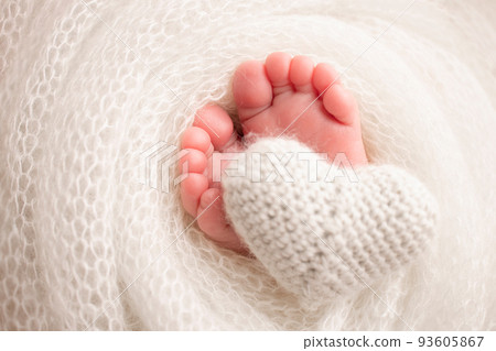 Knitted white heart in the legs of a baby. Soft feet of a new born in a white wool blanket. Close-up of toes, heels and feet of a newborn. Macro photography the tiny foot of a newborn baby. 93605867