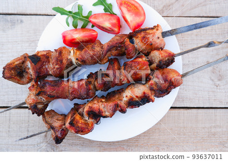 Juicy meat on skewers with tomatoes and herbs on a white plate on a wooden background. Kebab of meat is fried on the grill. 93637011