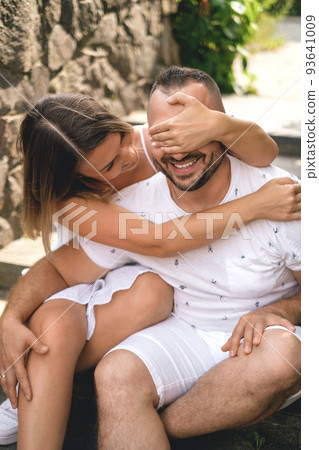 A young married couple in casual white clothes is sitting on the steps of a city street staircase and laughing merrily. The wife covers her husband's eyes with her hand 93641009