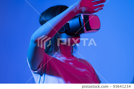 Excited brunette woman in white t-shirt getting experience using VR-headset glasses of virtual reality much gesticulating hands in neon light. 93641234