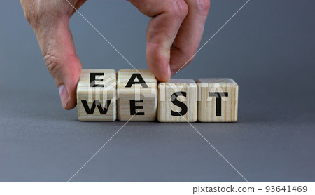 Hand turns a cube and changes the word 'east' to 'west', or vice versa. Beautiful grey background, copy space. Business concept. Hand turns a cube and changes the word 'east' to 'west', or vice versa. Beautiful grey background, copy space. Business concept. 93641469