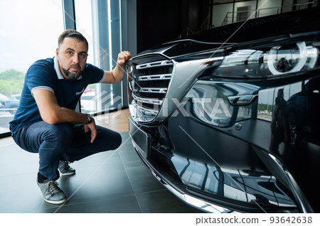 A happy Caucasian man sits near the hood of his new car in a car dealership. A happy Caucasian man sits near the hood of his new car in a car dealership. 93642638