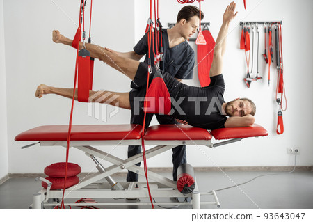 Rehabilitation specialist doing active treatment on suspension straps to a male patient Rehabilitation specialist doing active treatment on suspension straps to a male patient 93643047