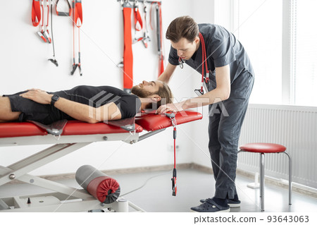 Rehabilitation specialist preparing for an active treatment on suspension straps with a male patient Rehabilitation specialist preparing for an active treatment on suspension straps with a male patient 93643063