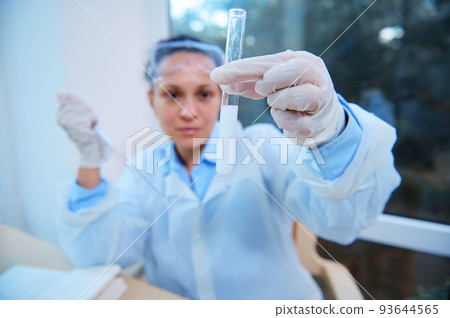 Detail: test tube with chemical preparation in the hands in medical protective gloves, of a blurred scientist, holding a graduated pipette and watching a chemical reaction going on in the test tube 93644565