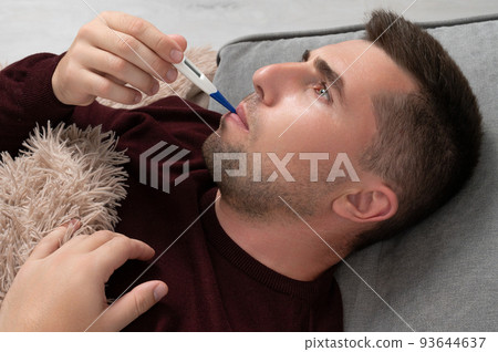 A man takes his temperature and holds his head with his hand because of a headache. A man takes his temperature and holds his head with his hand because of a headache. 93644637
