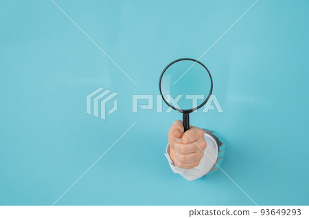 A woman's hand sticking out of a hole in a paper blue background holds a magnifying loupe. Copy space.  93649293
