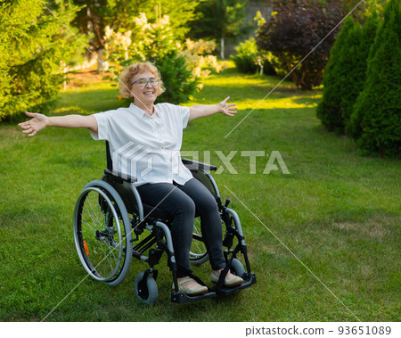 Happy old woman spread her arms to the sides while sitting in a wheelchair on a walk outdoors.  93651089
