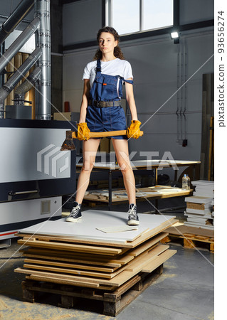 Woman in work clothes stands on a stack of chipboard slabs with an axe in her hands Woman in work clothes stands on a stack of chipboard slabs with an axe in her hands 93656274
