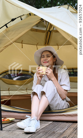 Relaxed young woman sitting inside camp tent and drinking hot coffee. Travel, adventure and vacation concept Relaxed young woman sitting inside camp tent and drinking hot coffee. Travel, adventure and vacation concept 93657024