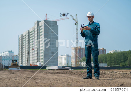 A man in a helmet and overalls controls a drone at a construction site. The builder carries out technical oversight. 93657974