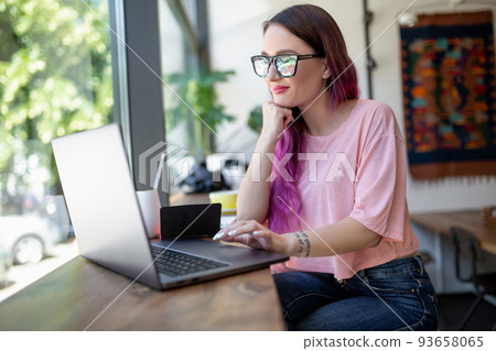 Side view of young businesswoman sitting at table in coffee shop. On table cup of coffee and laptop. In background white wall and window. 93658065