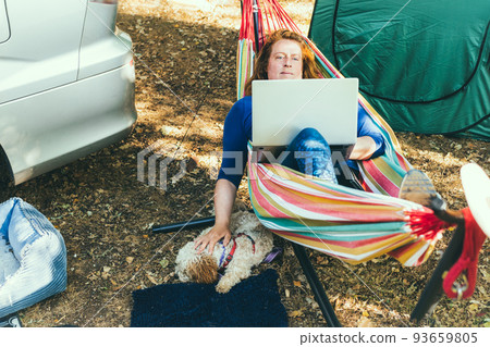 Adult woman working on laptop pc while lying in hammock, stroking cockapoo puppy near motorhome on camping trip. Female living on camper car with animals and travel the world. Remote worker people. Adult woman working on laptop pc while lying in hammock, stroking cockapoo puppy near motorhome on camping trip. Female living on camper car with animals and travel the world. Remote worker people. 93659805