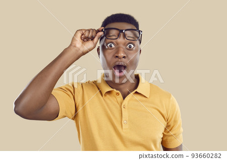 Young man opens his mouth, lifts glasses and looks at something with surprised face expression Young man opens his mouth, lifts glasses and looks at something with surprised face expression 93660282