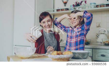 Smiling mother together with funny cute daughter taking selfie photo with smartphone camera making silly face while cooking at home in kitchen. Family, cook, and people concept 93660536
