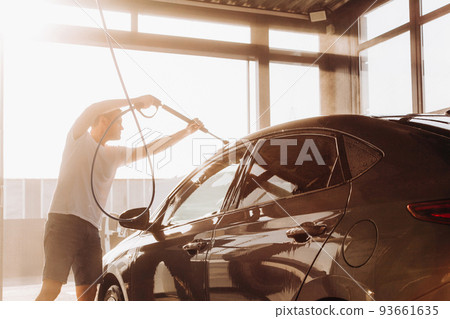 A man washes his car at a self-service car wash using a hose with pressurized water A man washes his car at a self-service car wash using a hose with pressurized water 93661635