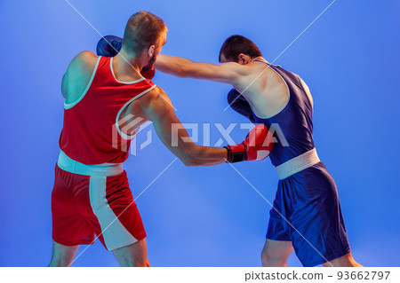 Left hook. Male professional boxers in red and blue sports uniform boxing isolated on blue background in neon light. Sport, skills, power, training, energy 93662797