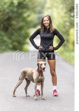 Young sporting woman and her Weimaraner dog stand together in the forest Young sporting woman and her Weimaraner dog stand together in the forest 93662860