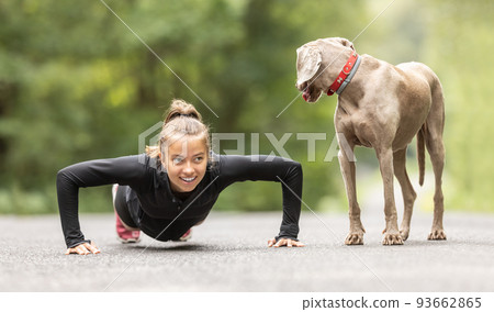 Young woman doing pushups outdoors smiles at her dog observing her Young woman doing pushups outdoors smiles at her dog observing her 93662865