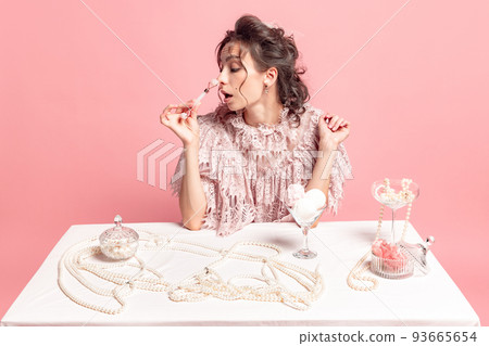 Beauty day. Creative image with young beautiful woman with retro style hairdo wearing medieval dress sitting at table with jewels isolated on pink background. 93665654