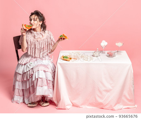Tasting pizza. Young happy beautiful girl with retro style hairdo wearing medieval dress sitting at served table isolated on pink background. Tasting pizza. Young happy beautiful girl with retro style hairdo wearing medieval dress sitting at served table isolated on pink background. 93665676