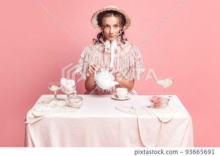 Tea ceremony. Young pretty woman with retro style hairdo in bonnet and vintage dress sitting at holiday table isolated on pink background. Tea ceremony. Young pretty woman with retro style hairdo in bonnet and vintage dress sitting at holiday table isolated on pink background. 93665691