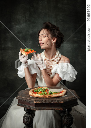 Tasting pizza. Vintage portrait of young beautiful girl in attire of medieval fashion style isolated on dark background. Comparison of eras concept, Tasting pizza. Vintage portrait of young beautiful girl in attire of medieval fashion style isolated on dark background. Comparison of eras concept, 93665692