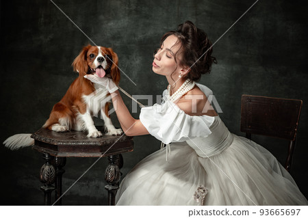 Beloved pet. Young beautiful woman in image of medieval person in renaissance style dress with little cute dog isolated on dark background. Beloved pet. Young beautiful woman in image of medieval person in renaissance style dress with little cute dog isolated on dark background. 93665697