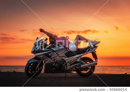 Freedom. Sexy young woman posing on the motorcycle. Golden sunset and motorbike on the background. Side view. Concept of World Motorcyclist Day and moto trips 93666014