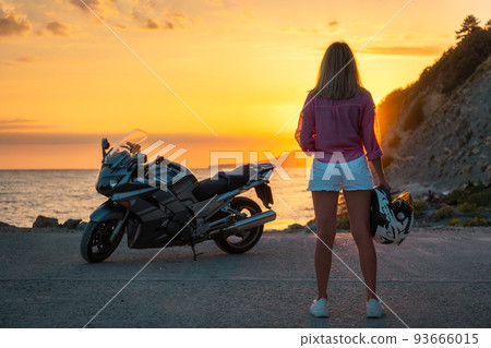 Young woman posing at the beach holds a helmet. Golden sunset and motorbike on the background. Back view. Concept of World Motorcyclist Day and moto trips 93666015