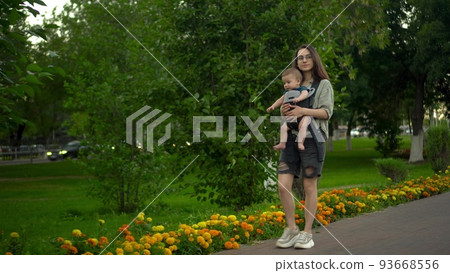 A young woman walks along the alley in the park with her son. The girl in glasses walks. Newborn son in a kangaroo-backpack. 93668556