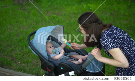 A young mother shows a rattle toy to a child. The baby lies in a stroller in the park. A woman with glasses takes care of her son. A young mother shows a rattle toy to a child. The baby lies in a stroller in the park. A woman with glasses takes care of her son. 93668565