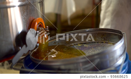 The beekeeper opens the tap with honey. Honey is filtered through a sieve after extraction. The beekeeper opens the tap with honey. Honey is filtered through a sieve after extraction. 93668625