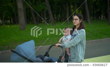 A young mother walks with a six-month-old son carrying him in her arms. A woman with glasses carries a baby and pushes a stroller in the park. 93668627