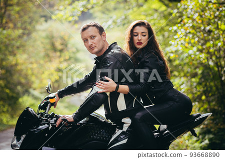 Handsome young man and beautiful young girl, couple sitting on a motorcycle in black leather clothes, close up, hugging, in nature, outdoors, looking at camera Handsome young man and beautiful young girl, couple sitting on a motorcycle in black leather clothes, close up, hugging, in nature, outdoors, looking at camera 93668890