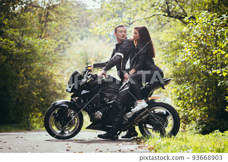 Handsome young man and beautiful young girl, couple sitting on a motorcycle, standing near a motorcycle in black leather clothes, hugging, in nature, on the street Handsome young man and beautiful young girl, couple sitting on a motorcycle, standing near a motorcycle in black leather clothes, hugging, in nature, on the street 93668903